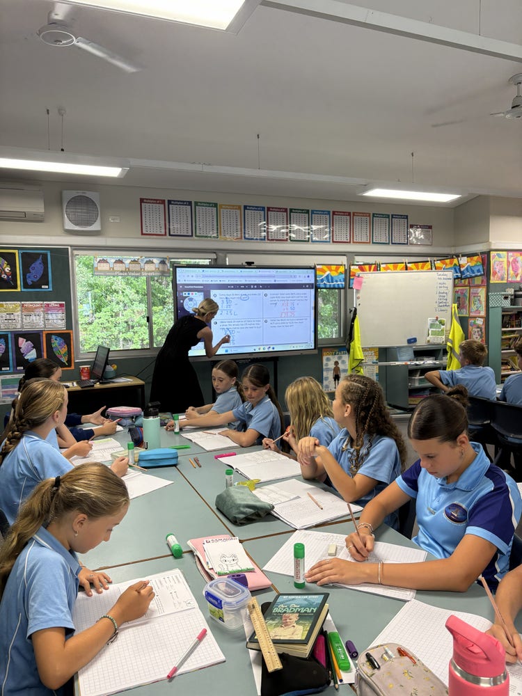 students sitting at the table watching the teacher write on the whiteboard
